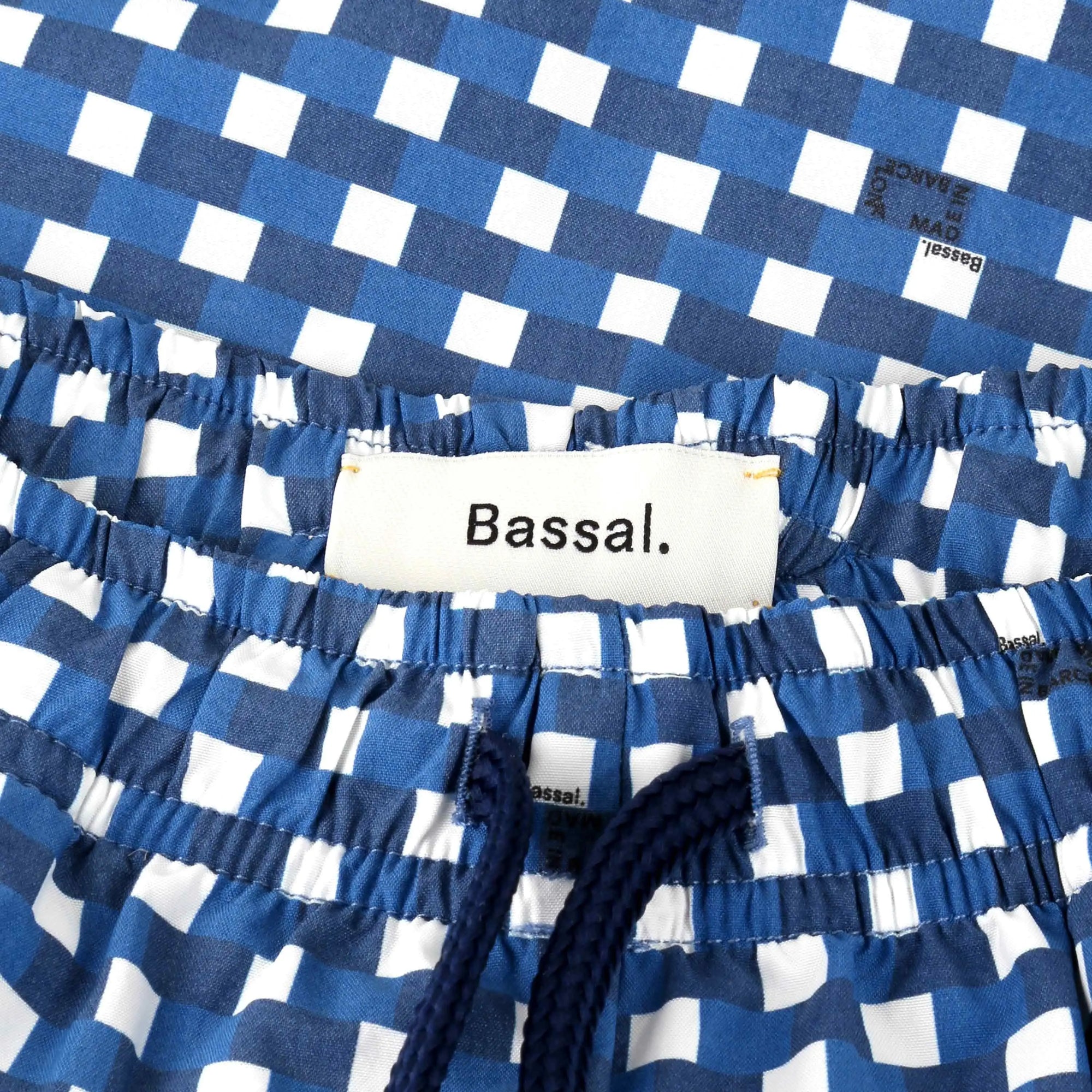 A pair of Paris Blue Swimwear is neatly folded inside an orange box with white edges. Attached to the swimwear are a white tag with the brand name "Bassal." and a card. The box lid, partially visible, also bears the brand name "Bassalstore," likely from their Barcelona store.