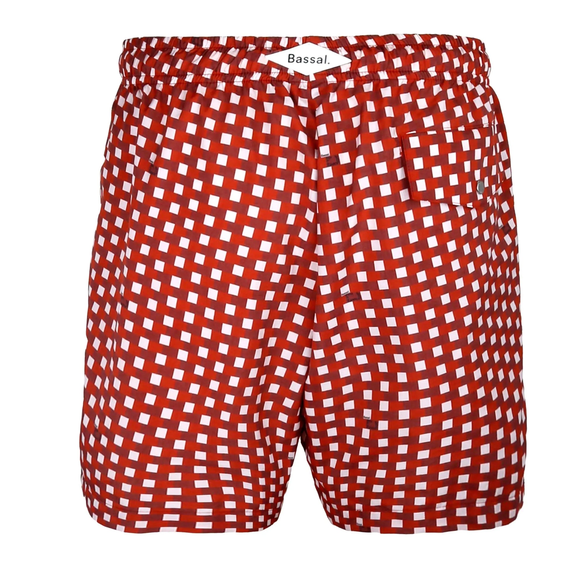 A pair of red and white checkered Paris Red Swimwear with an orange drawstring waistband are neatly folded inside an open box. A tag attached to the swimwear reads "Bassal." The box, showcased by BassalStore, and accompanying items are displayed in a clean, minimalist style against a white background.