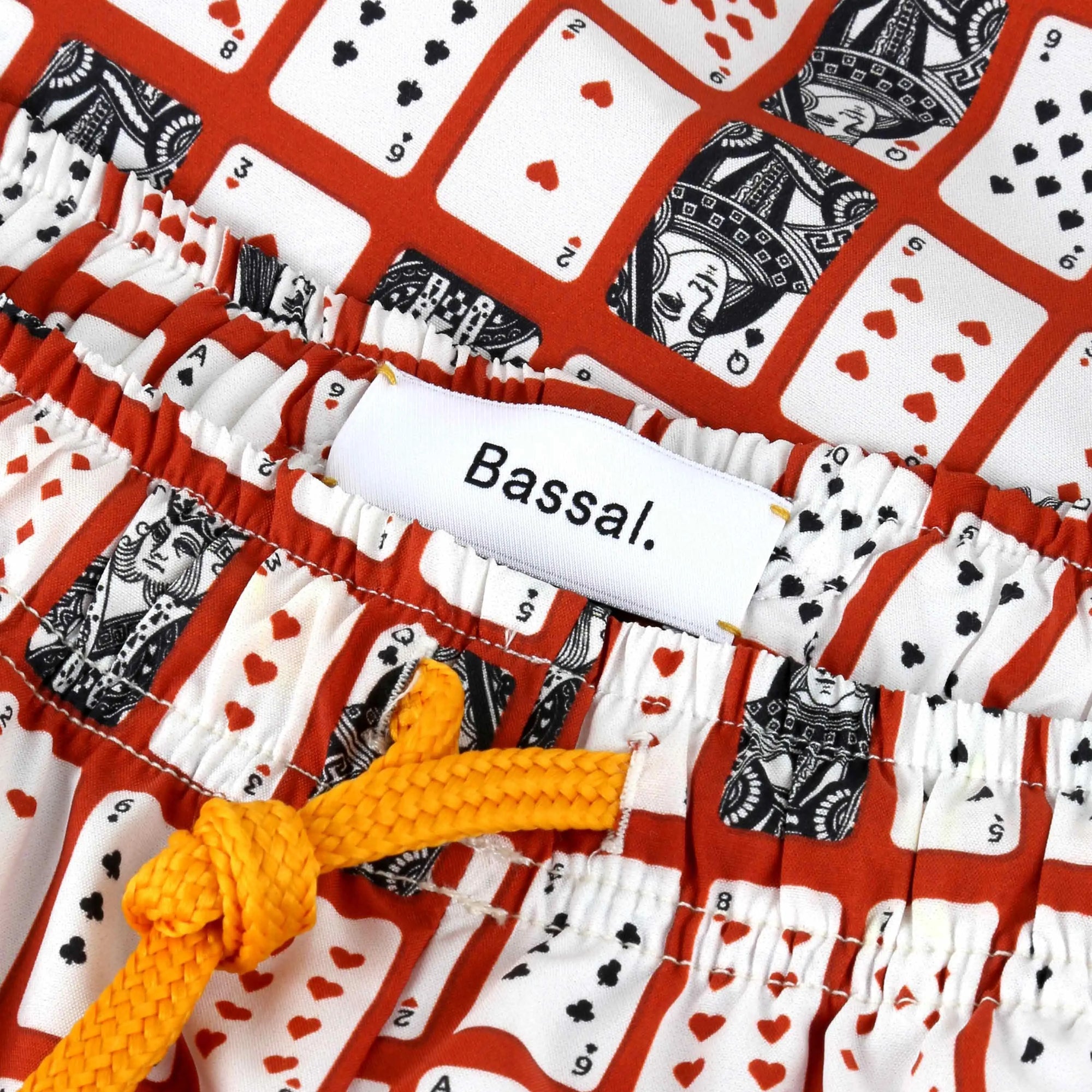 A small white box with a lid partially removed reveals a fabric bag inside. The bag, boasting a colorful playing card pattern and an orange drawstring, is from the Bassal. store in Barcelona. Attached to the bag are two tags—one blue and one white—with text including "Poker Swimwear".