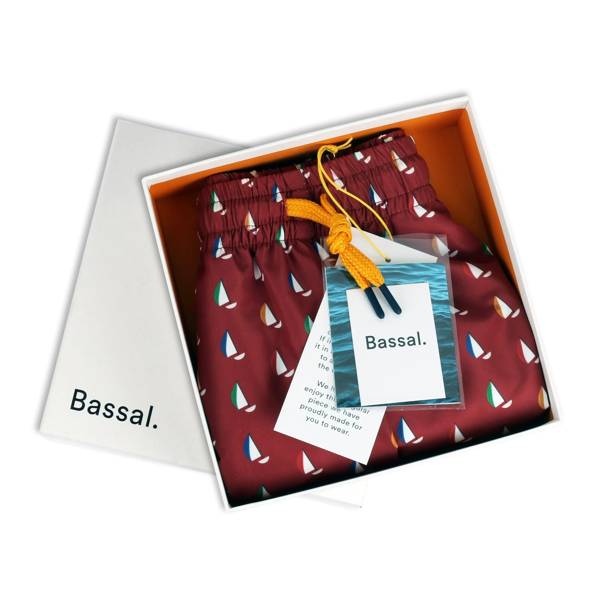 An open white box with the brand name "Bassal." on the lid from the renowned Bassalstore in Barcelona contains Regata Swimwear adorned with colorful sailboat patterns. The swimwear is neatly tied with a yellow drawstring and comes with a tag featuring the same brand name and an image of a sailboat.