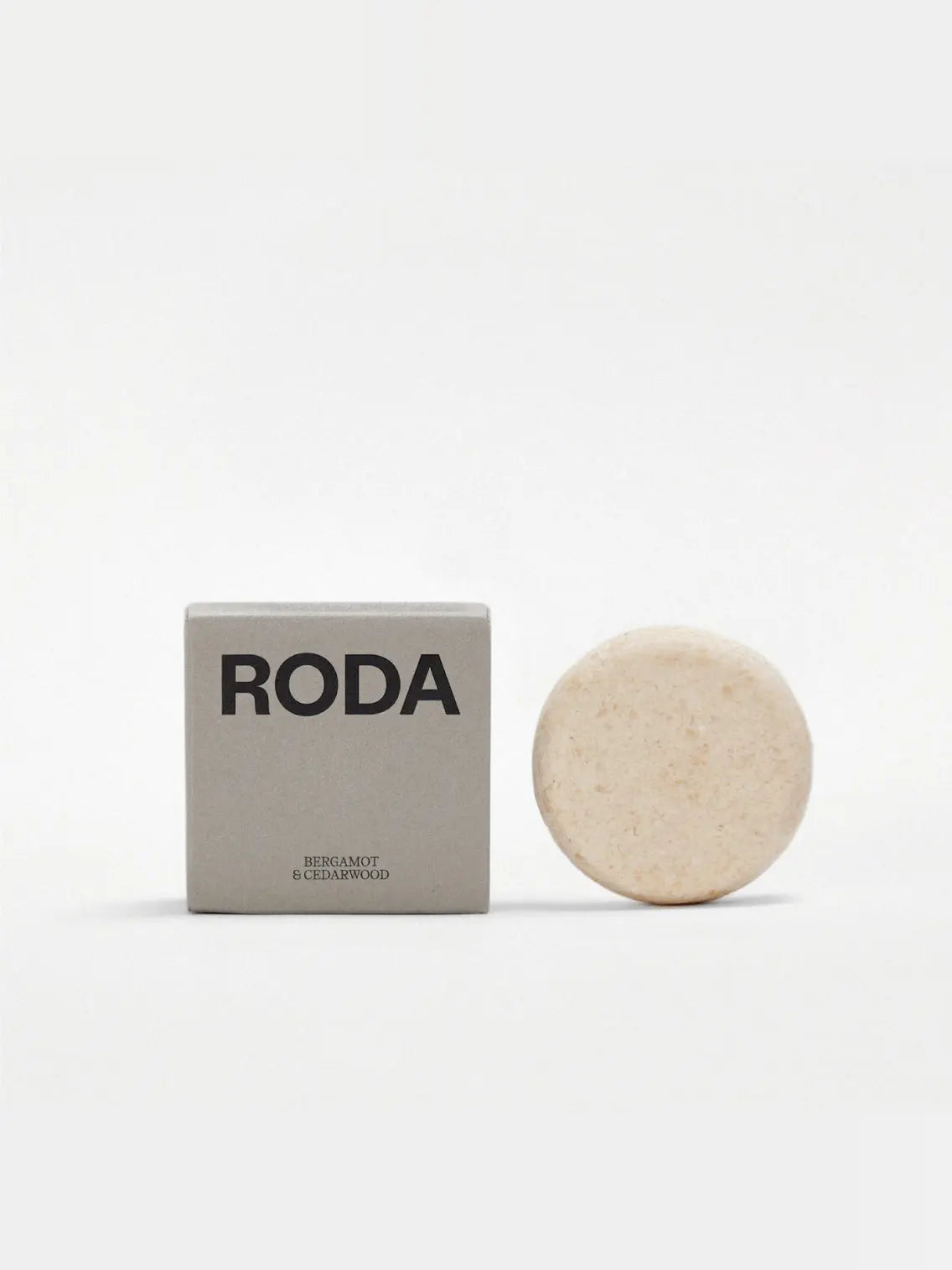 A round, beige shampoo bar stands next to a rectangular gray box labeled "Roda". The box has black text and indicates the bar's scent as "Bergamot & Cedarwood" on a white background. This elegant product is available at your local Barcelona store, Bassalstore.
