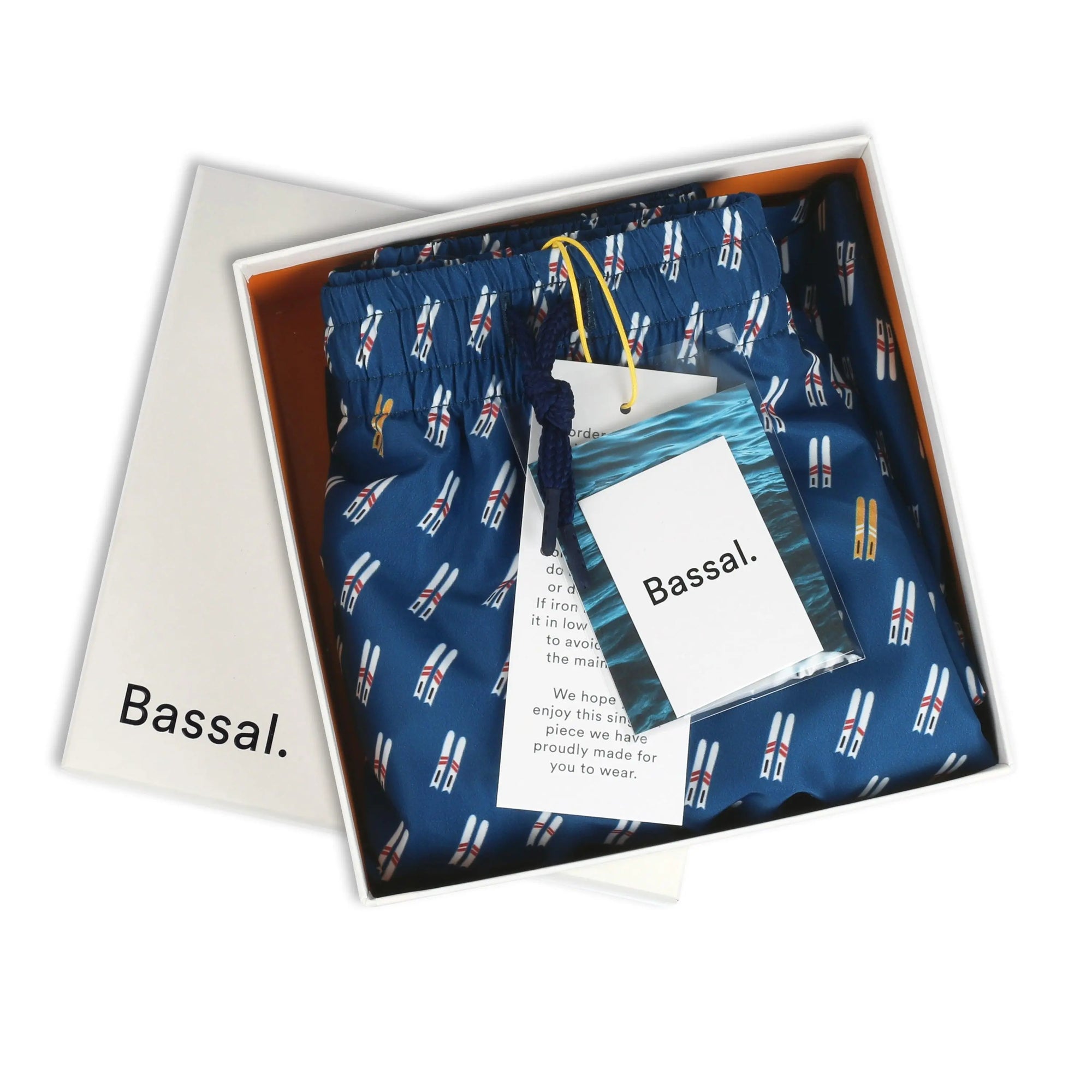 A box of Ski Swimwear men's swim trunks with a blue base color and a pattern of small surfboards. The open box, branded with the name "Bassal," hints at the quality you'd expect from Barcelona's premier store. A tag and an additional label attached to the trunks are visible.