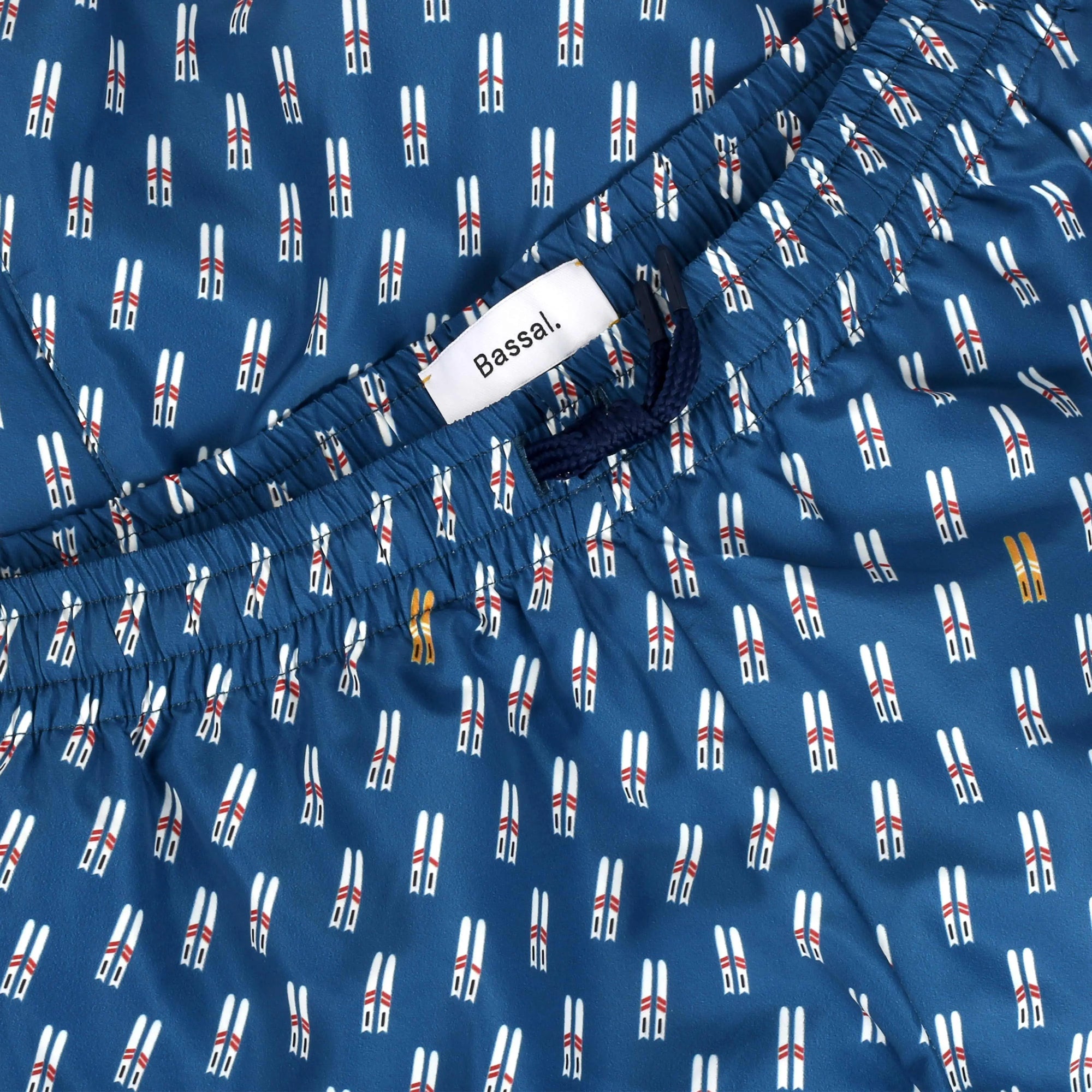 A box of Ski Swimwear men's swim trunks with a blue base color and a pattern of small surfboards. The open box, branded with the name "Bassal," hints at the quality you'd expect from Barcelona's premier store. A tag and an additional label attached to the trunks are visible.