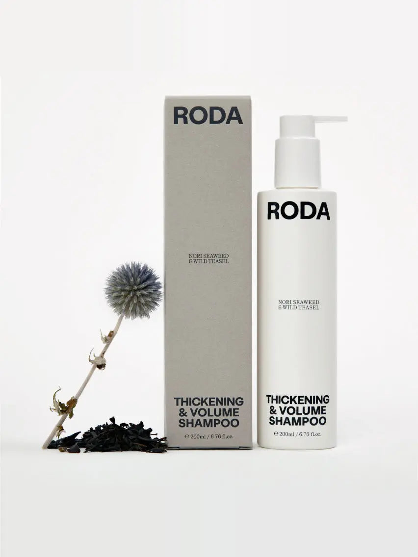 A bottle of Roda Thickening & Volume Shampoo is placed alongside its gray box. The shampoo bottle is white with a pump dispenser and black text. Nearby, there is a sprig of dried flowers and a small pile of dark seaweed. Available at Bassalstore in Barcelona, the bottle contains 200ml (6.7 fl. oz.).