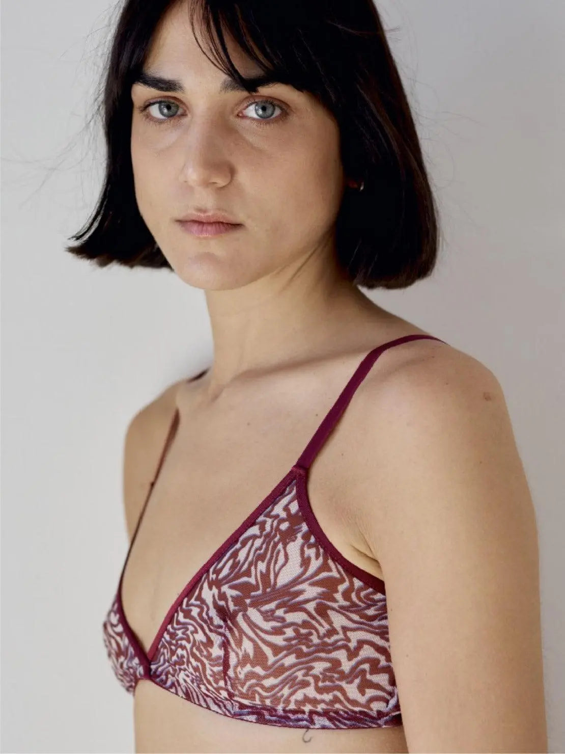 A woman with shoulder-length dark hair and bangs, wearing a patterned Triangle Bra with thin straps, looks into the camera with a neutral expression. The bra, showcasing a red and white intricate design from Interiors in Barcelona, stands out against the plain, light-colored background.