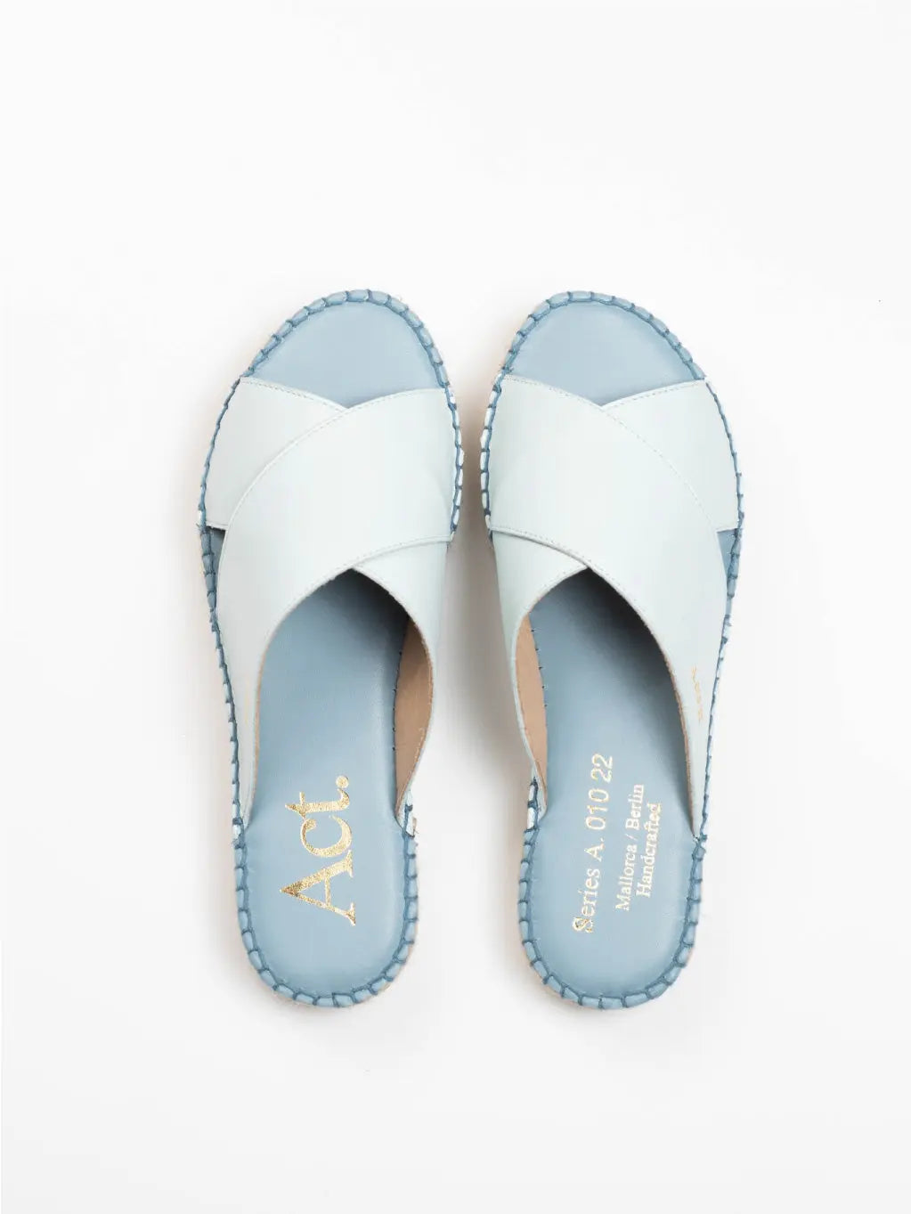 A pair of Uccle Light Blue Espadrilles with crisscross straps and exposed toes. The insole features gold text on the left sandal, and the stitching around the edges is in a matching light blue color. The espadrilles from Act Series are placed on a white background.