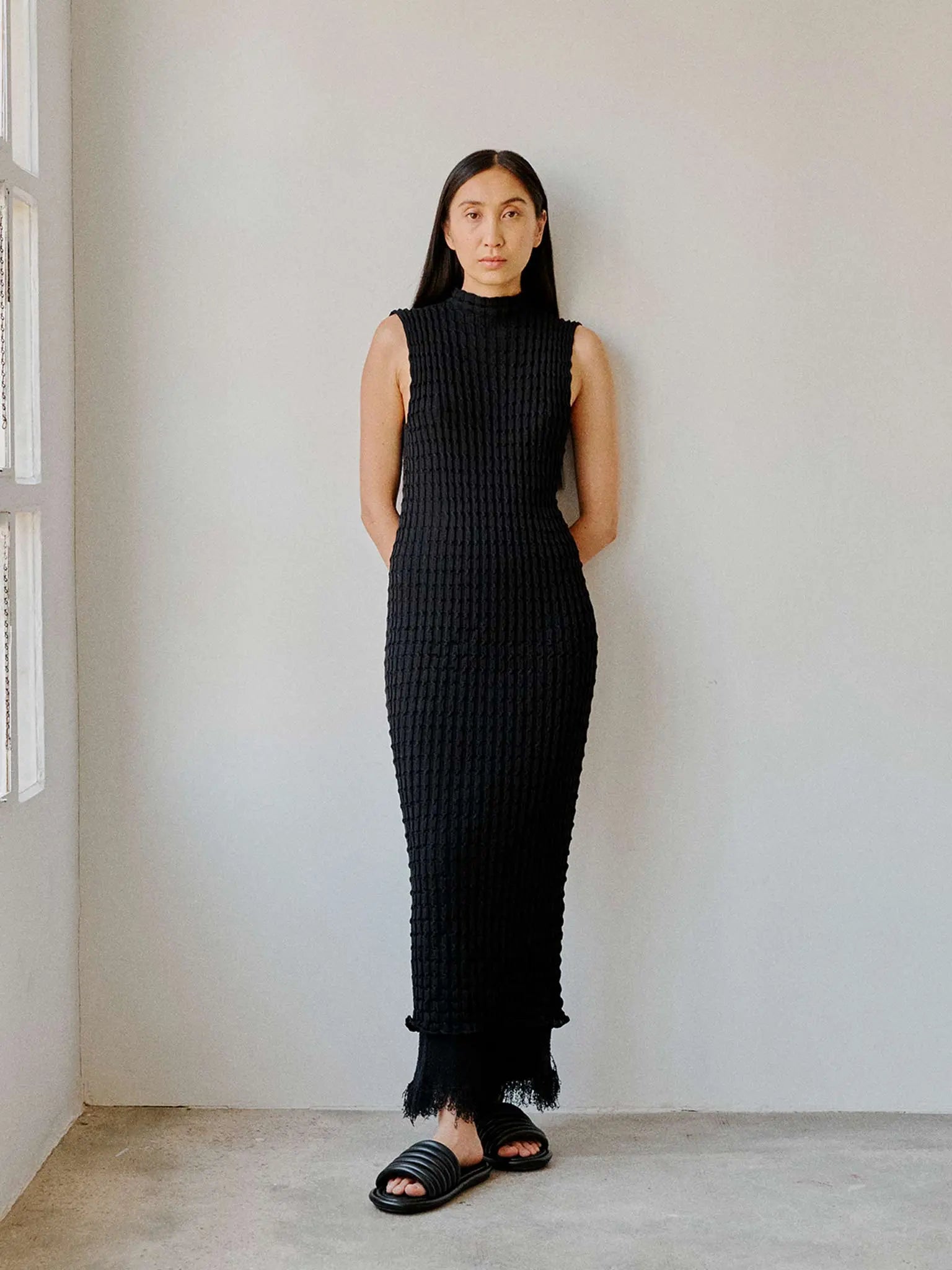 Image of the Udon Dress Ink by Rus, a black, sleeveless, ribbed-knit dress with a high neckline and a fitted silhouette. The dress extends to mid-calf length, showcasing a sleek, elegant design. Available at Bassalstore in Barcelona, the hemline has a slight scalloped edge.