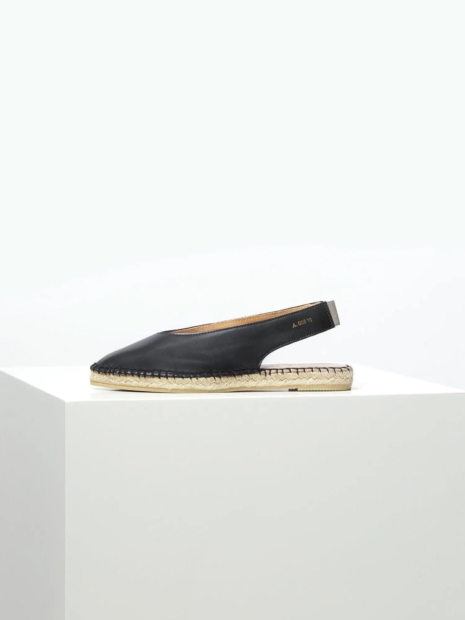 A single Ulvik Black Leather Espadrille with an open back and a beige woven jute sole is displayed on a white geometric pedestal against a plain white background. The shoe, marked "Act Series" near the heel, epitomizes the chic style found in Barcelona's trendiest stores like Bassalstore.