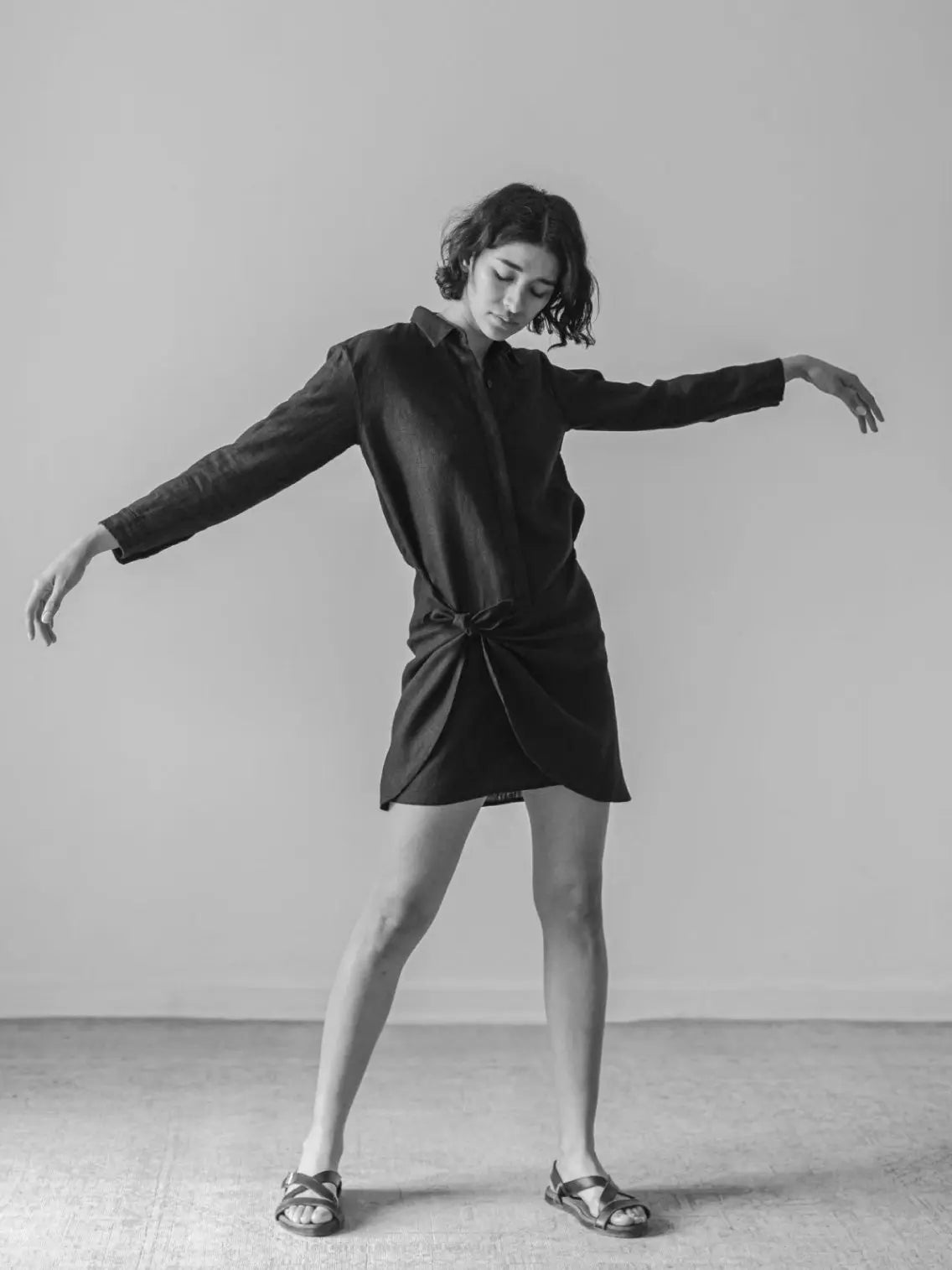 A black and white photo of a person standing on one leg with eyes closed and arms gracefully extended. They are wearing the Vane Longsleeve Dress by Zii Ropa with long sleeves and sandals, embodying an elegance reminiscent of old Barcelona. The background is plain and minimalistic.