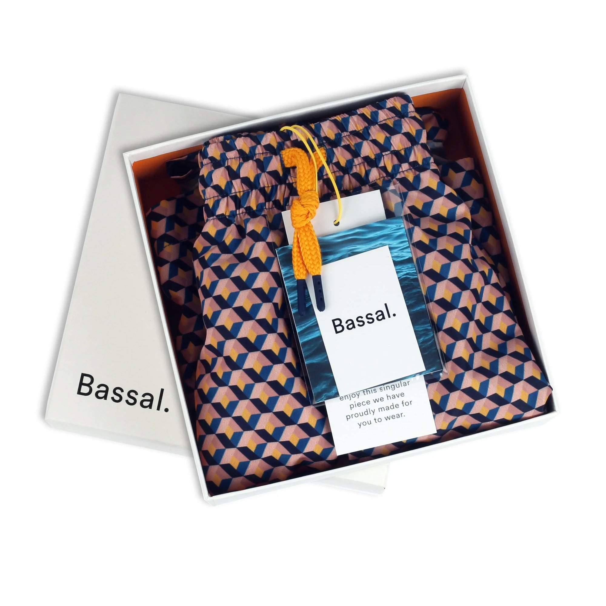 Open white box from Bassalstore with "Bassal." written on the lid, containing a folded pair of Vee Swimwear in orange, blue, and white. The swimwear has a tag attached, and the lid has another tag with a message inside. A stylish piece straight from Barcelona.