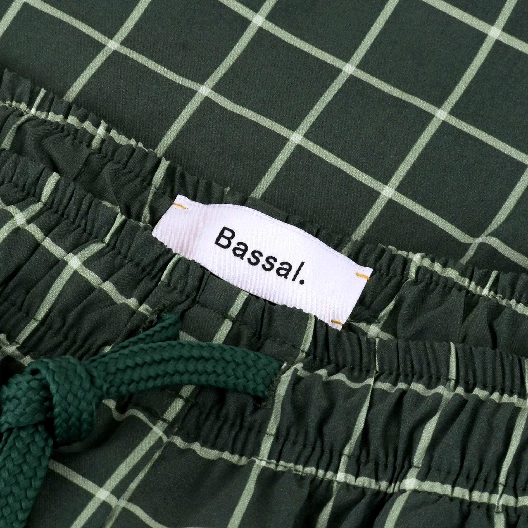 A pair of Verd Swimwear with a white grid pattern neatly folded inside a white box, along with a white product tag bearing the brand name "Bassal." The box lid, placed beside it, also displays the "Bassal" logo and hints at its exclusive availability at bassalstore.