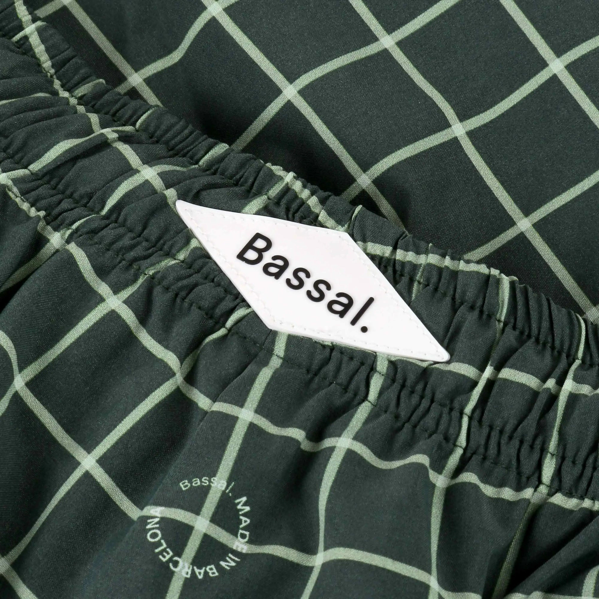 A pair of Verd Swimwear with a white grid pattern neatly folded inside a white box, along with a white product tag bearing the brand name "Bassal." The box lid, placed beside it, also displays the "Bassal" logo and hints at its exclusive availability at bassalstore.