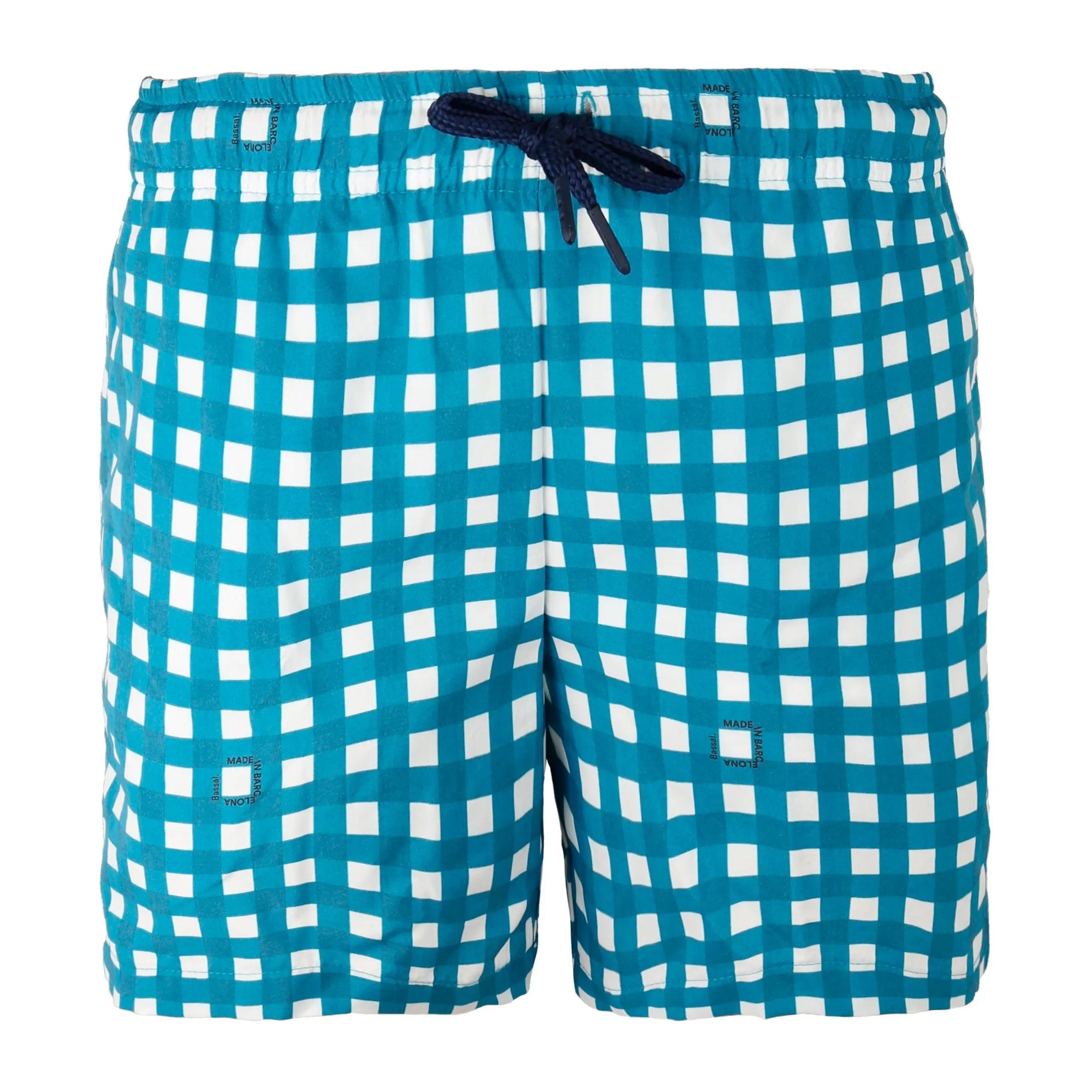 A pair of blue and white checkered Vichy Kids Swimwear with a navy drawstring is neatly folded inside an open orange box. Attached to the swimwear is a white tag with the brand name "Bassal." The box, featuring the "Bassalstore" logo, is placed on top of another box in a chic store in Barcelona.