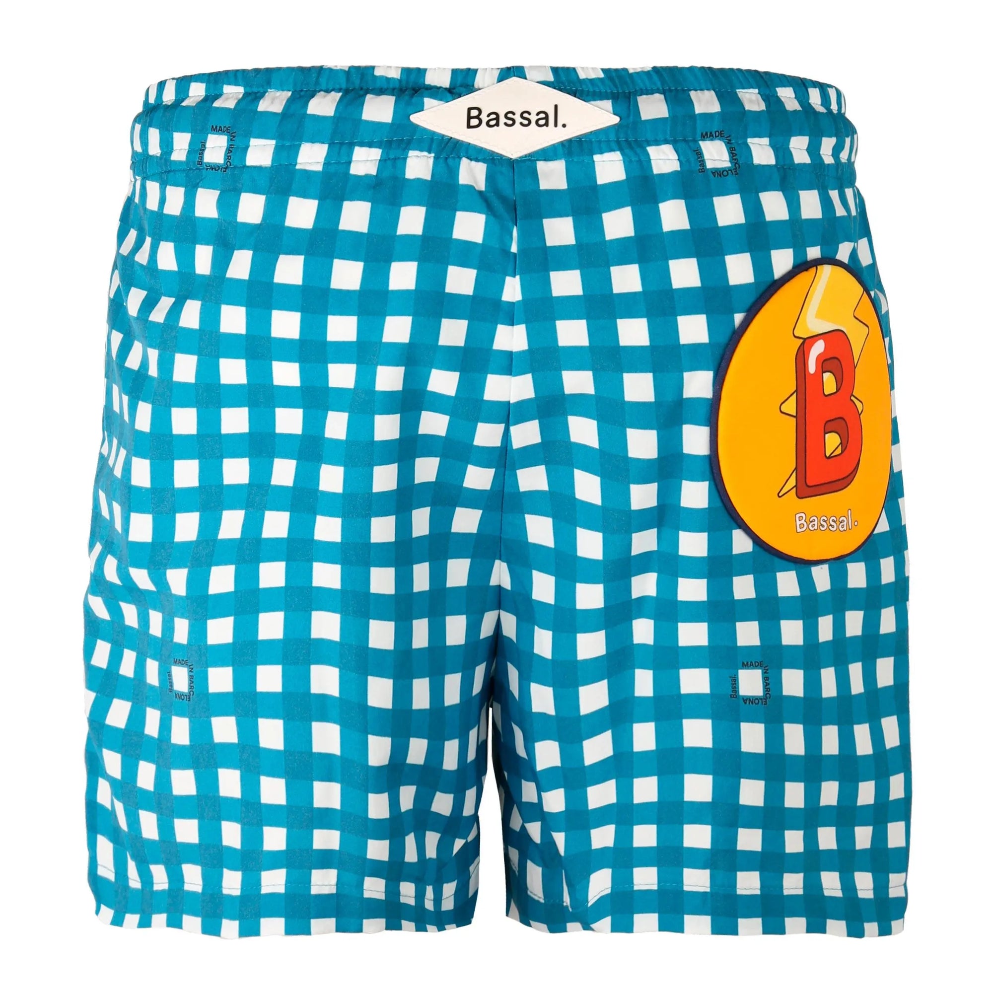 A pair of blue and white checkered Vichy Kids Swimwear with a navy drawstring is neatly folded inside an open orange box. Attached to the swimwear is a white tag with the brand name "Bassal." The box, featuring the "Bassalstore" logo, is placed on top of another box in a chic store in Barcelona.