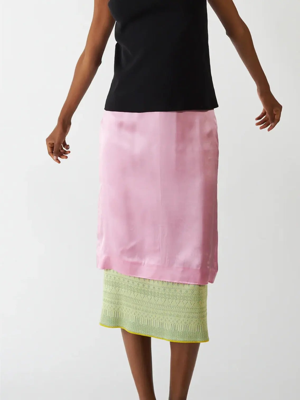 A person is wearing a black sleeveless top paired with the Vivi Skirt Candy by Bielo, a knee-length pink silk skirt layered over a pastel green crocheted skirt that extends slightly below the pink skirt. This chic ensemble, available at Bassalstore in Barcelona, stands out beautifully against the plain white background.