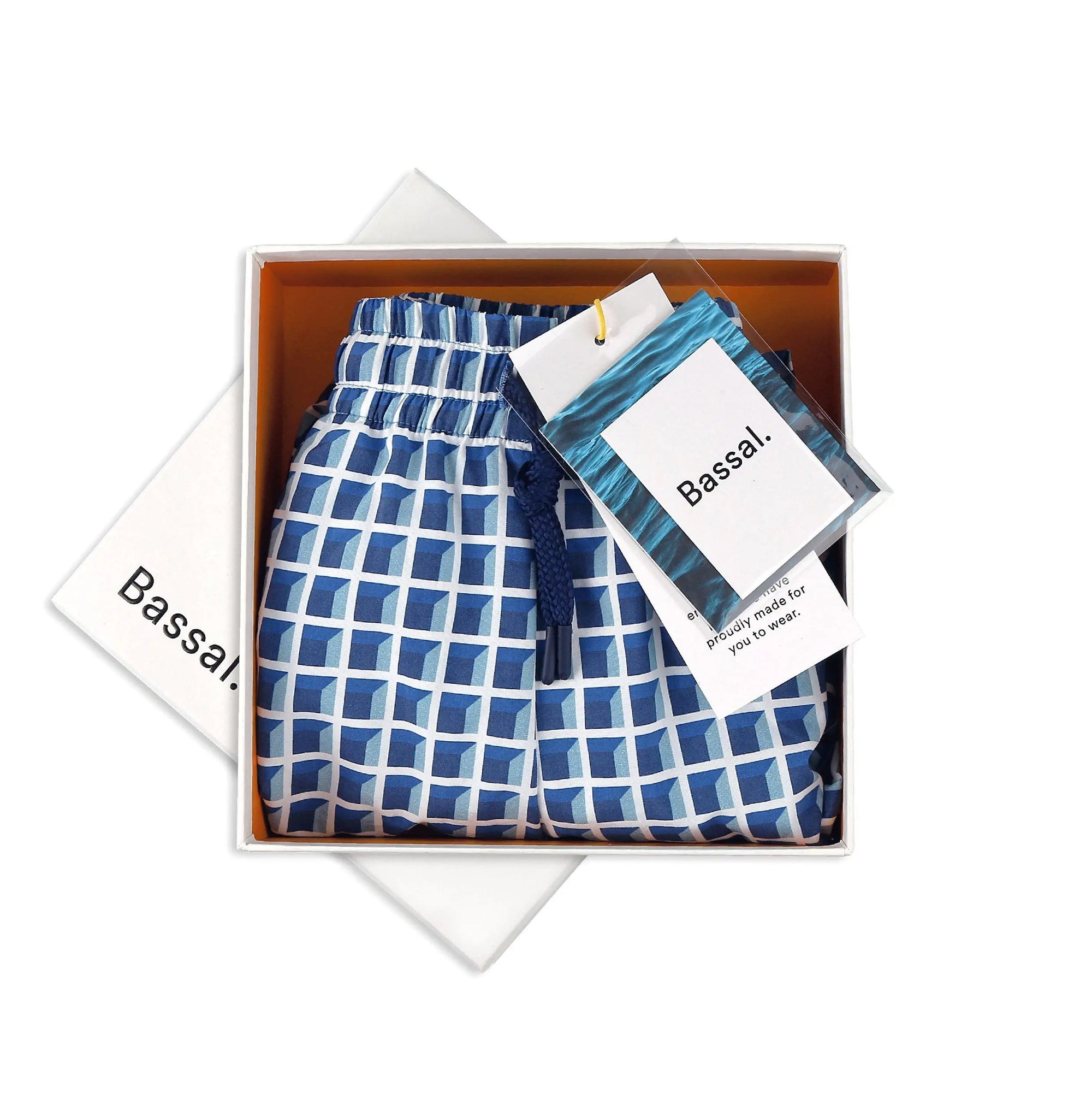 A pair of Window Blue Swimwear are neatly folded inside an orange gift box with the brand name "Bassal" on the lid and tags. The swimwear features an elastic waistband with a blue drawstring. Another white box with "BassalStore" is partially visible underneath, hinting at a chic store experience in Barcelona.