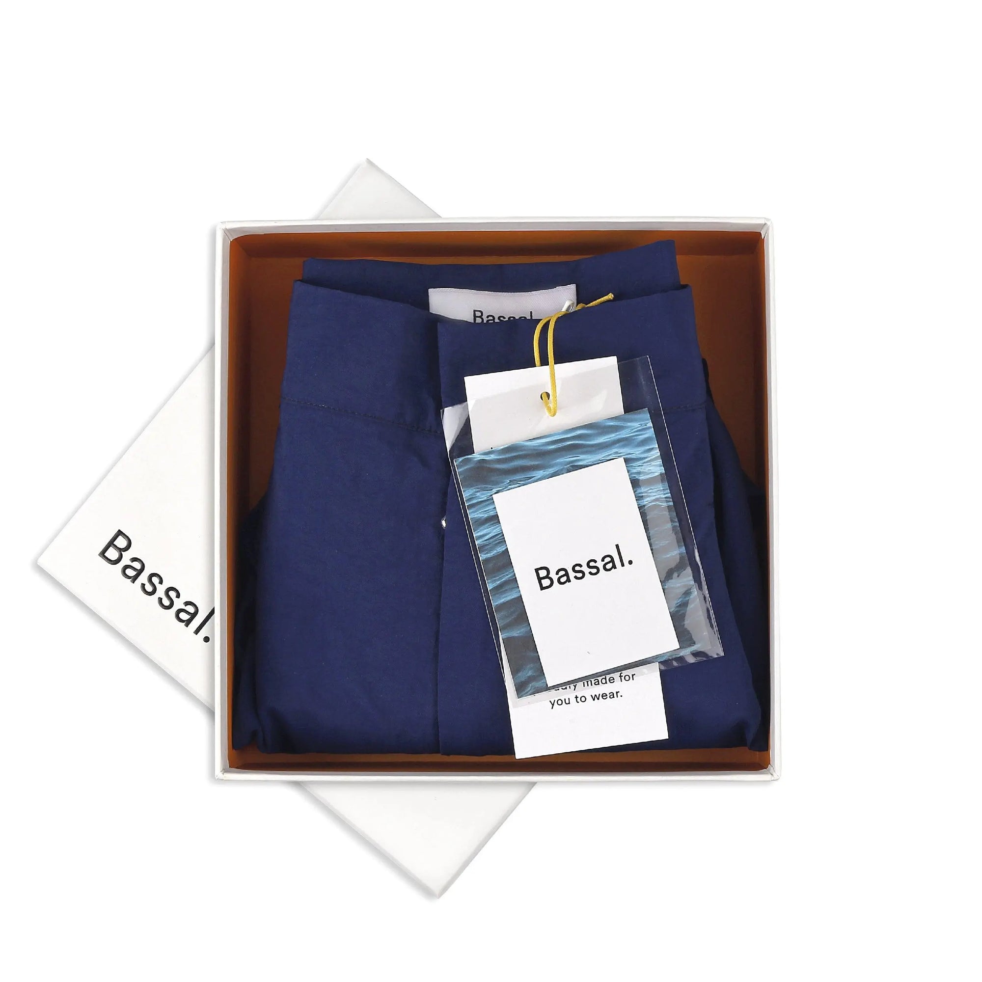 A neatly folded dark blue bermuda swimwear is placed in a white box with a brown interior. The box lid, slightly off to the side, and a tag on the swimwear both feature the brand name "Bassal." A small plastic packet is attached to the tag. Discover this elegant piece exclusively at BassalStore, your go-to for Barcelona style.